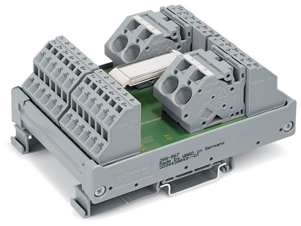 Ein grauer elektrischer Anschluss mit mehreren Anschlüssen für Leitungen, der auf einer grünen Leiterplatte montiert ist und mit technischen Details beschriftet ist.