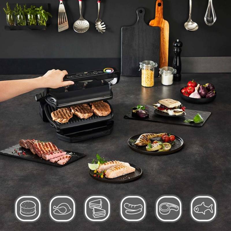 Eine Person, die verschiedene Fleisch- und Gemüsesorten auf einem Elektrogrill in einer modernen Küche grillt, wobei Küchenutensilien zu sehen sind.