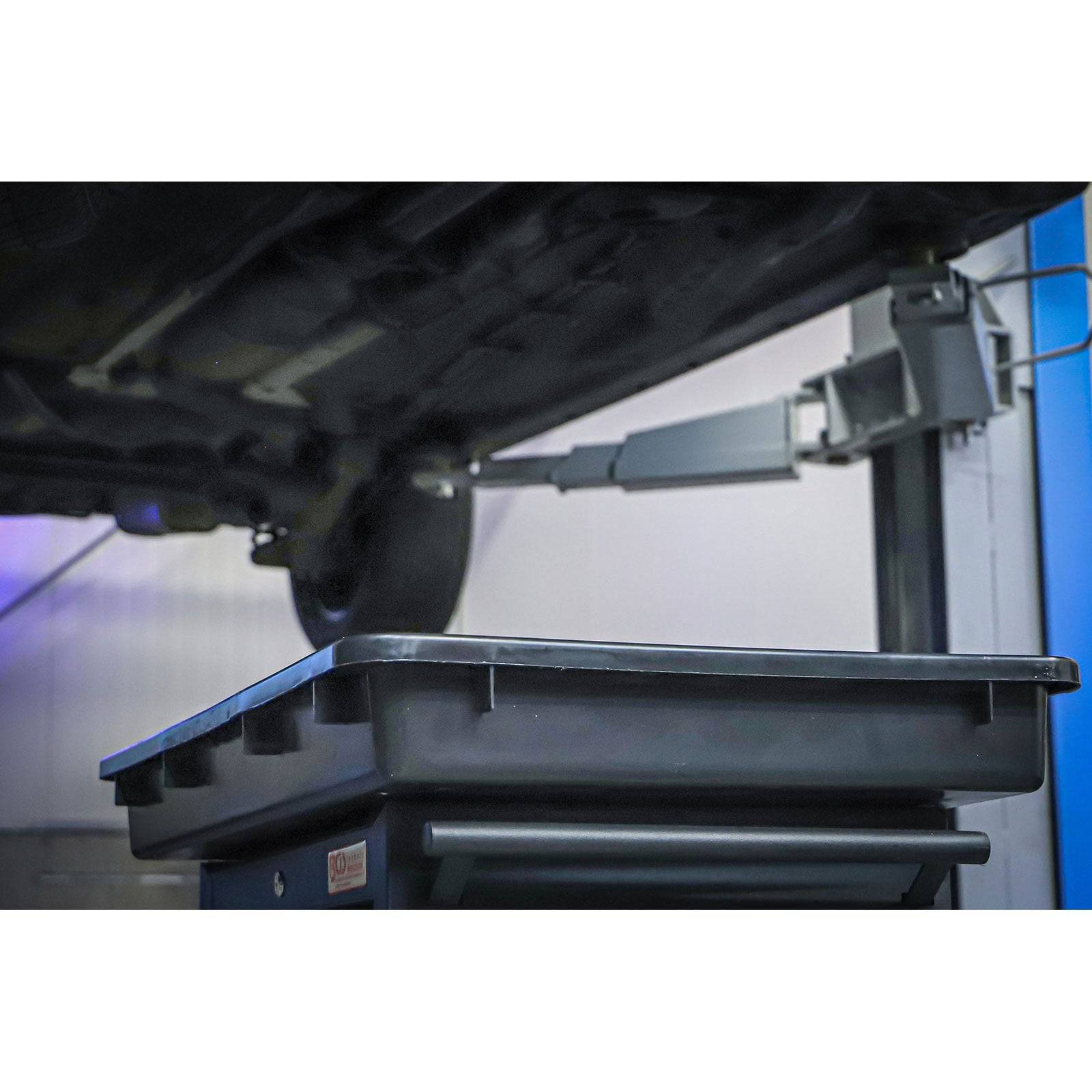 Ein Auto wird in einer Autowerkstatt auf einer Hebebühne angehoben, wobei eine große Auffangwanne darunter platziert ist, um auslaufende Flüssigkeiten aufzufangen.