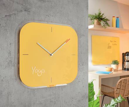 Gelbe quadratische Wanduhr mit der Aufschrift „Yoga" hängt an einer Betonwand neben einem hellen Heim-Arbeitsbereich mit Regalen und einem Schreibtisch.
