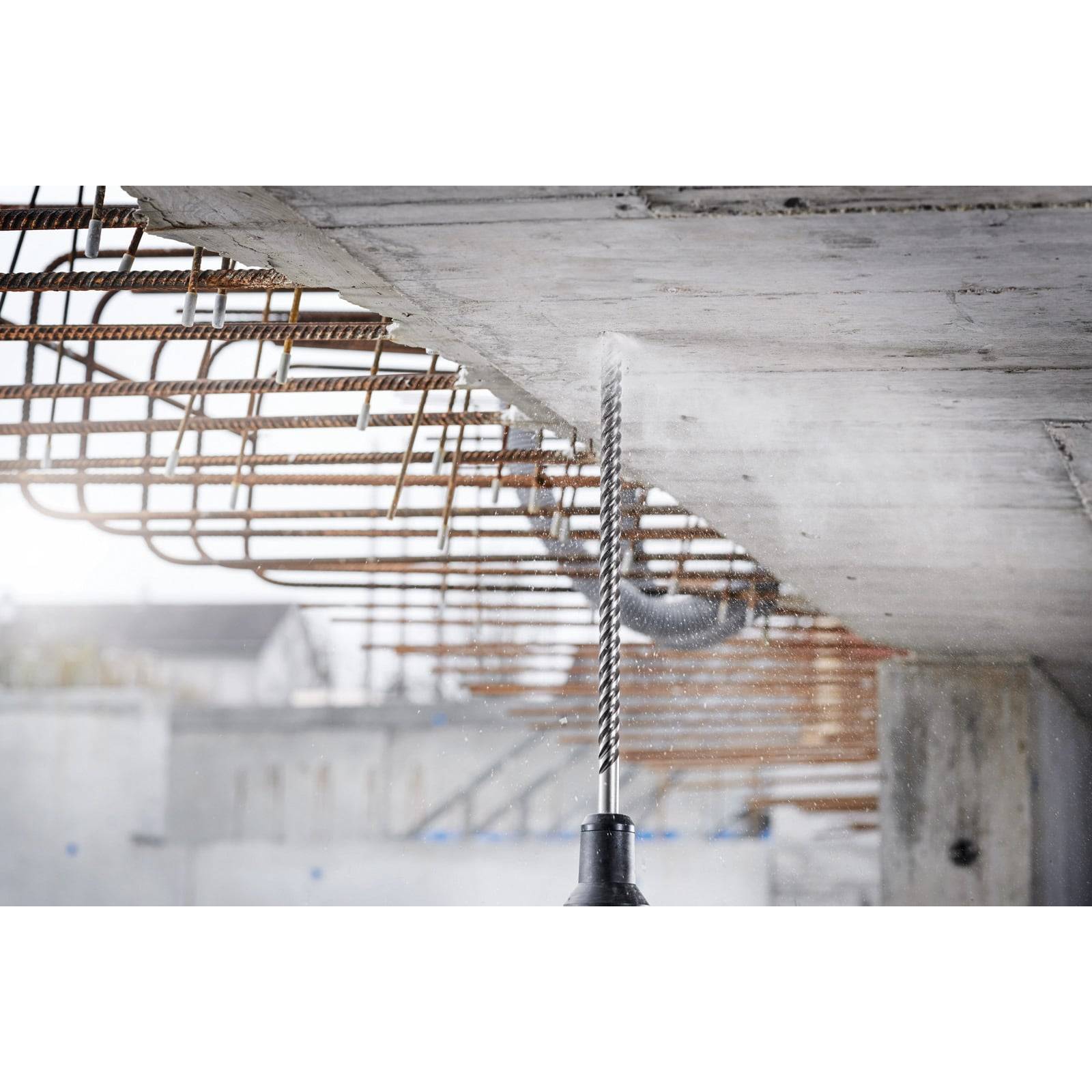 Ein Bohrer durchbohrt eine Betondecke, umgeben von freiliegenden Stahlträgern, auf einer Baustelle und signalisiert aktive Bauarbeiten.