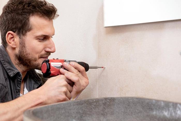 Eine Person, die einen roten Akkuschrauber verwendet, um ein Loch in eine hellefarbene geflieste Wand neben einem Waschbecken zu bohren, was auf eine Heimwerkeraufgabe hindeutet.