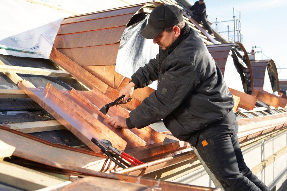Ein Arbeiter auf einem Dach repariert oder installiert Holzpaneele mit Hammer und Nägeln inmitten von verstreuten Dachmaterialien.