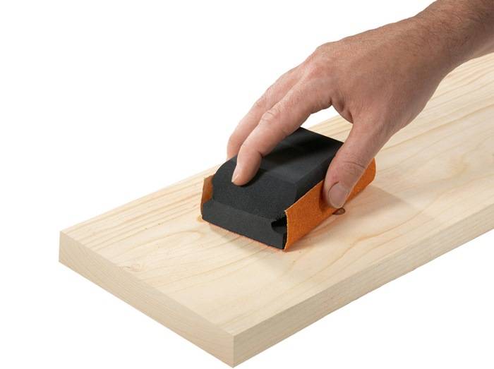 Eine Hand, die ein Holzbrett mit einem Schleifklotz und schwarzem Schleifpapier schleift und dabei den Prozess des Glättens einer Oberfläche demonstriert.