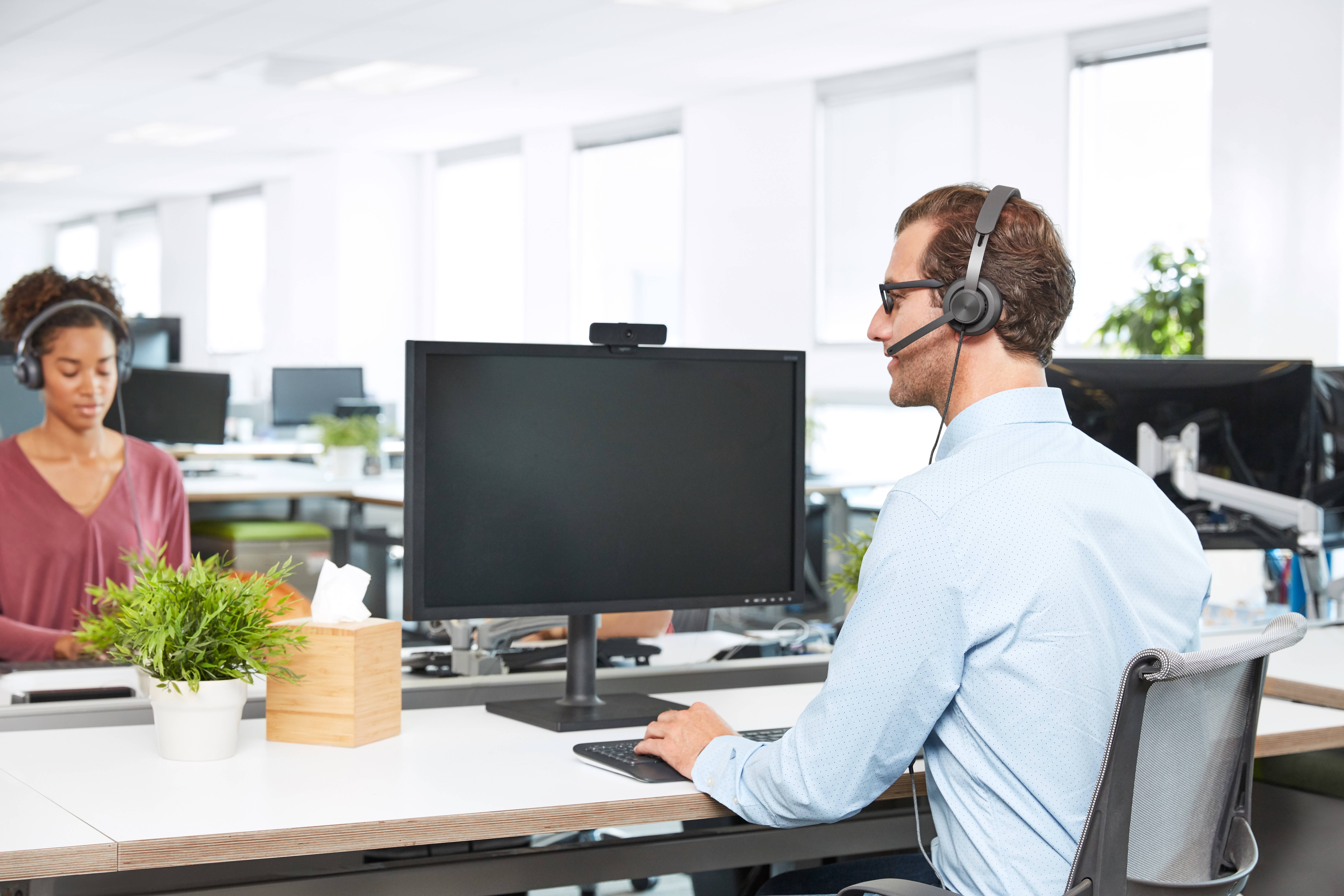 Kundendienstmitarbeiter mit Headset, der an einem Computer in einem hellen Büro arbeitet, während er mit einem Kollegen über den Schreibtisch hinweg spricht.