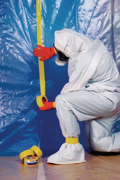 Eine Person in einem vollständigen Schutzanzug klebt blaue Plastikfolie an eine Wand, möglicherweise als Teil eines Eindämmungs- oder Reinigungsvorhabens.