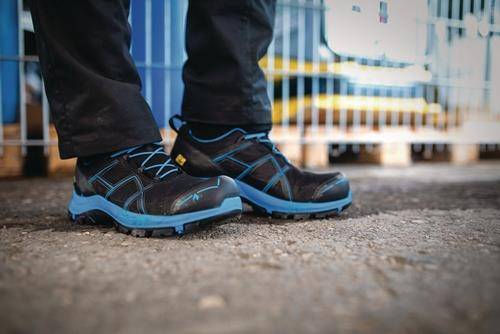 Eine Person trägt schwarze und blaue Sicherheitsarbeitsschuhe und steht auf einer Betonoberfläche, während im Hintergrund industrielle Ausrüstung unscharf zu sehen ist.