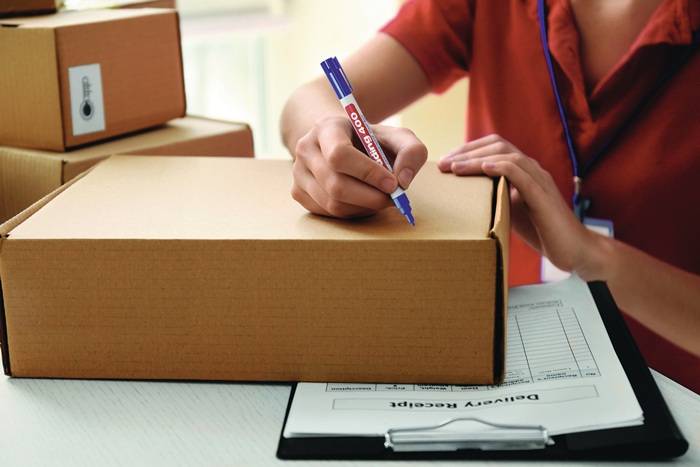 Eine Person schreibt mit einem blauen Stift auf einen Karton, neben einem Klemmbrett mit einem Lieferschein.