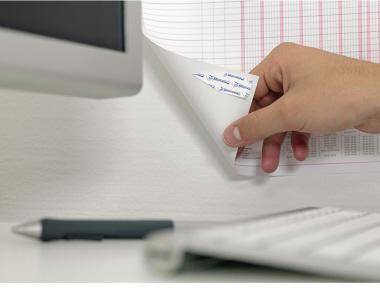 Eine Hand legt ein gefaltetes Papier in einen an der Wand montierten Dokumentenhalter neben einem Computermonitor, mit einem Stift und einem Notizbuch auf dem Schreibtisch.
