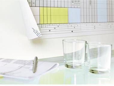 Schreibtisch mit Dokumenten und zwei leeren Gläsern vor einer Wandtafel oder einer Tabelle mit hervorgehobenen Spalten.