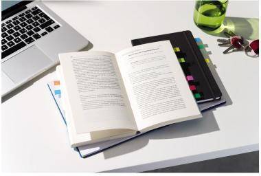 Offenes Buch auf einem weißen Schreibtisch neben einem Laptop, Notizbüchern, farbigen Registerkarten, einem grünen Getränk und Schlüsseln, was auf Studium oder Büroarbeit hindeutet.
