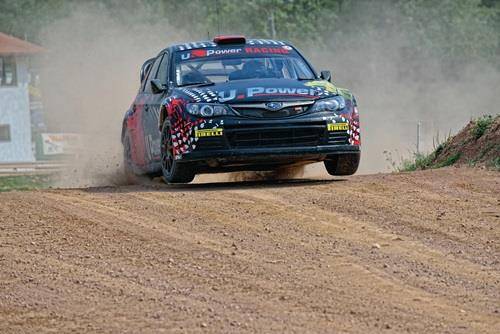 Ein Rallyeauto rast auf einer Schotterpiste und wirbelt Staub auf. Das Fahrzeug ist mit Sponsorenlogos bedeckt. Dichter Baumbestand säumt den Hintergrund.