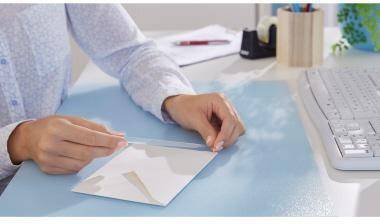 Eine Person fertigt einen weißen Umschlag handwerklich an einem Schreibtisch an, wo Papier, eine Tastatur und Büromaterialien in der Nähe sind.