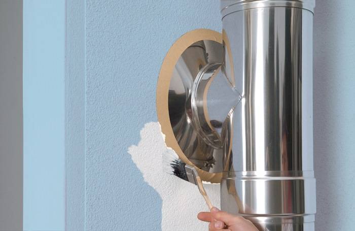 Eine Hand, die eine Wand mit einer Rolle hellblau anstreicht und dabei um ein glänzendes Metallrohr herum arbeitet, das aus der Wand hervorsteht.