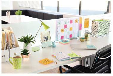 Heller Bürotisch mit Laptop, Lampe, Pflanze, Kaffeetasse, Notizbüchern und einer farbcodierten Wandplanungstafel in einem modernen Arbeitsbereich.