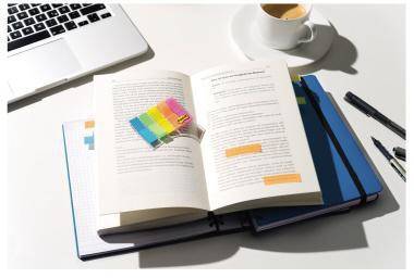 Offenes Buch mit farbigen Haftnotizen auf einem Schreibtisch neben einem Laptop, einer Kaffeetasse, Notizbüchern und Stiften.