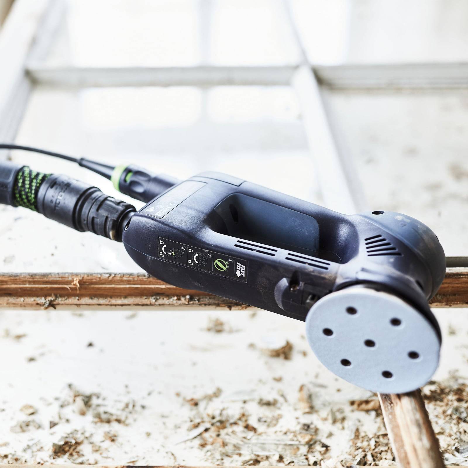 Eine handgehaltene elektrische Schleifmaschine liegt auf einer Holzoberfläche mit Sägemehl, neben einem Fensterrahmen, was auf Hausrenovierung oder Schreinerarbeiten hindeutet.