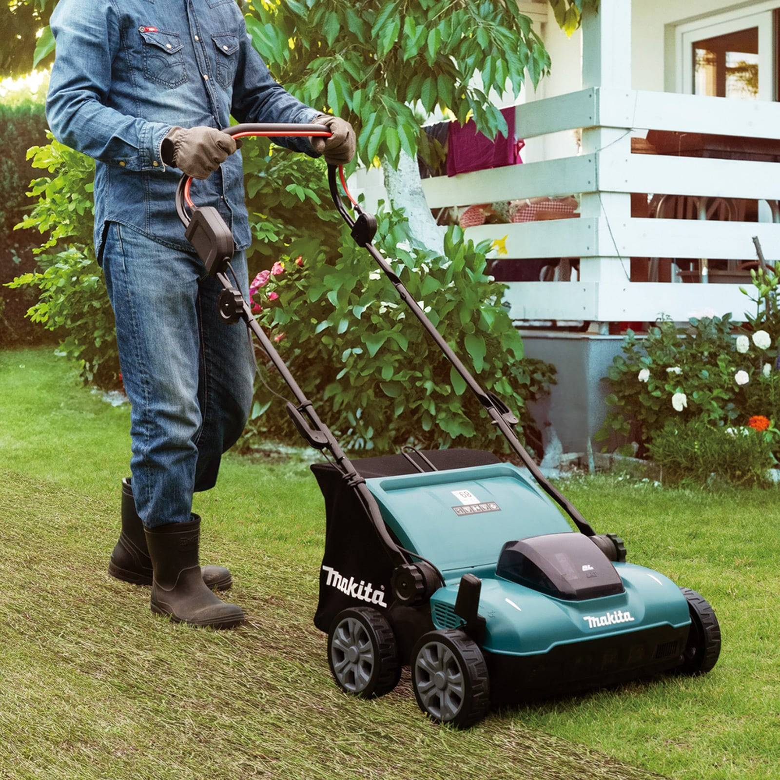 Eine Person in Jeankleidung verwendet einen Makita-Rasenmäher, um Gras in einem üppigen Garten mit blühenden Pflanzen und einem weißen Haus im Hintergrund zu schneiden.