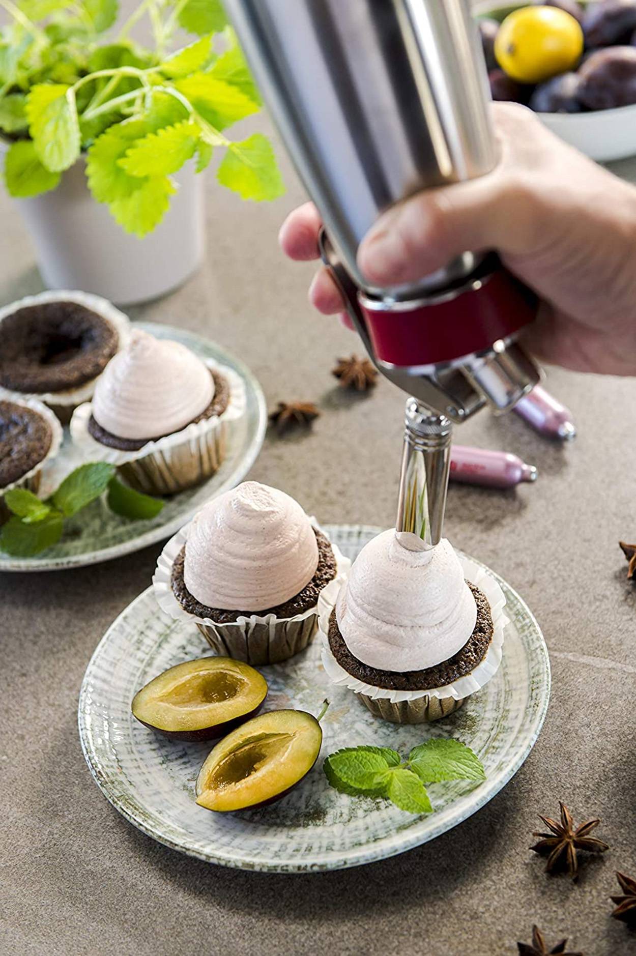 Mit der Hand Creme-Topping auf Schokoladencupcakes auf einem Teller spritzen, mit geschnittenen Früchten und Minzgarnitur auf einer Küchencounterplatte.