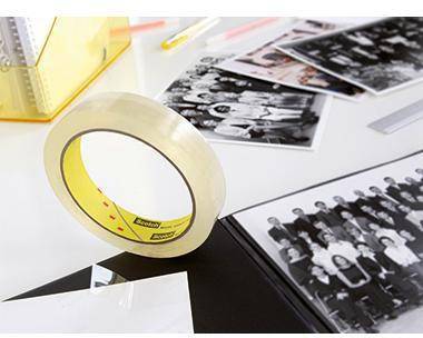Rolle durchsichtiges Klebeband auf einem Schreibtisch neben verstreuten Schwarz-Weiß-Fotodrucken und Papieren.