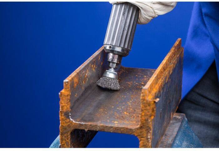 Ein Arbeiter verwendet eine rotierende Drahtbürste, um Rost von einem Metallträger zu entfernen. Das Metall zeigt Anzeichen von Rost und deutet auf einen laufenden Reinigungsprozess hin.