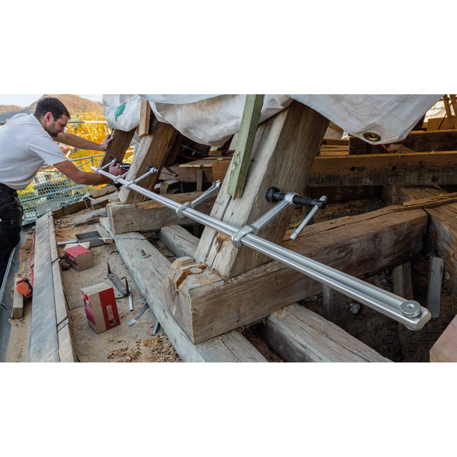 Ein Arbeiter justiert eine Metallstützstruktur auf einem Holzdach und ist möglicherweise an einem Bau- oder Restaurierungsprojekt beteiligt.