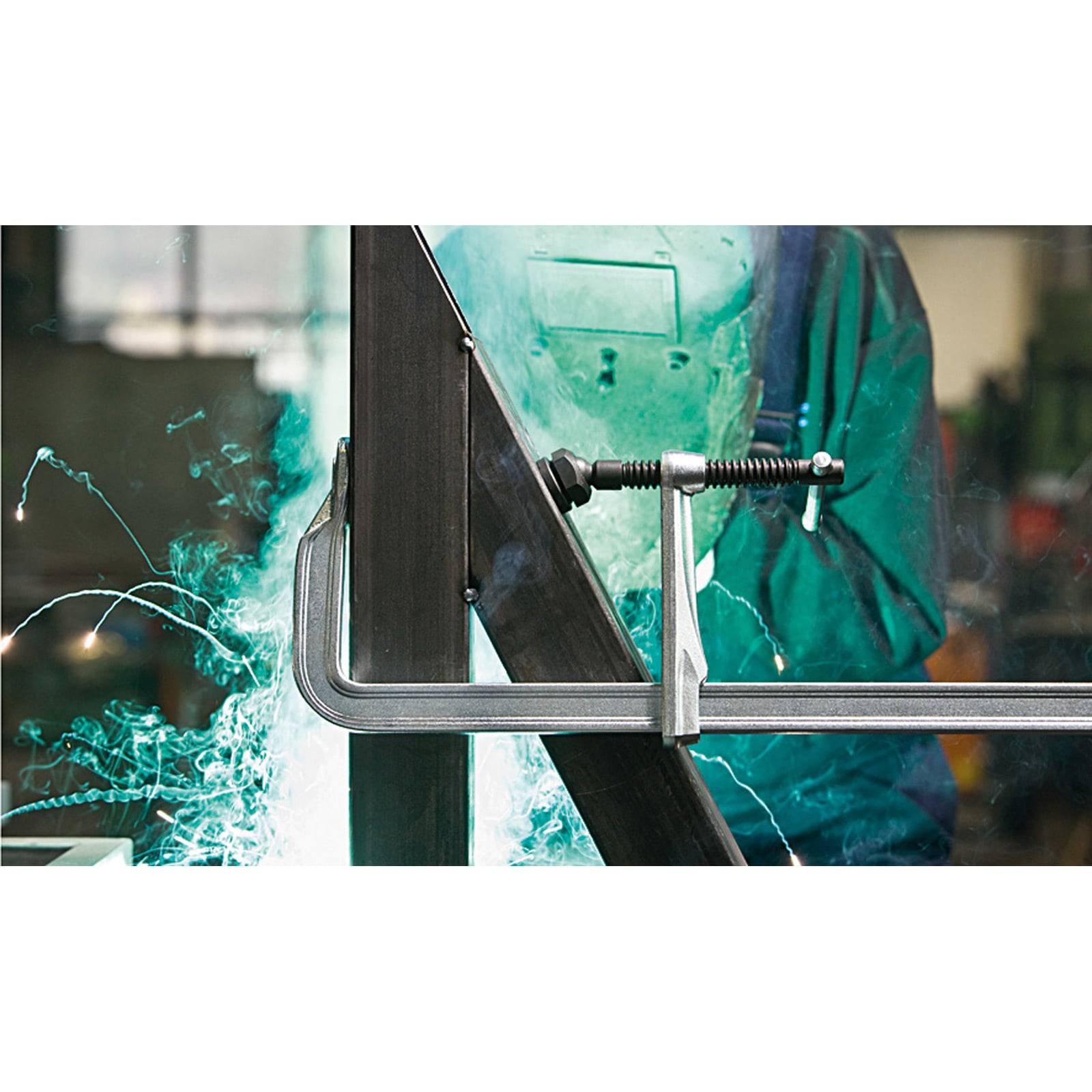 Eine Person in Schutzausrüstung schweißt Metall mit einer C-Schraube. Helle Funken sind sichtbar und heben den Schweißvorgang in einer Werkstattumgebung hervor.