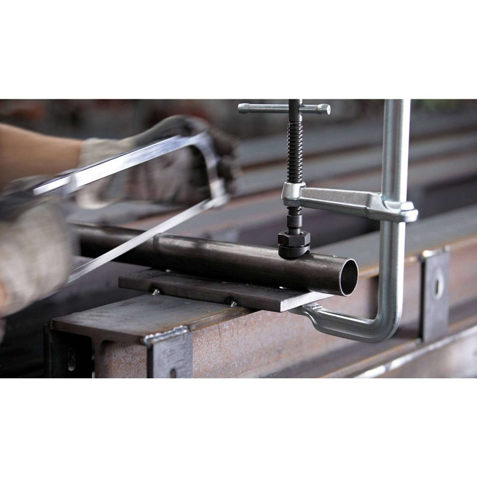 Eine Person verwendet eine Metallsäge (Hacksaw), um ein mit einer Klemme auf einem Stahlträger befestigtes Metallrohr zu sägen und damit einen Metallbearbeitungsprozess zu demonstrieren.