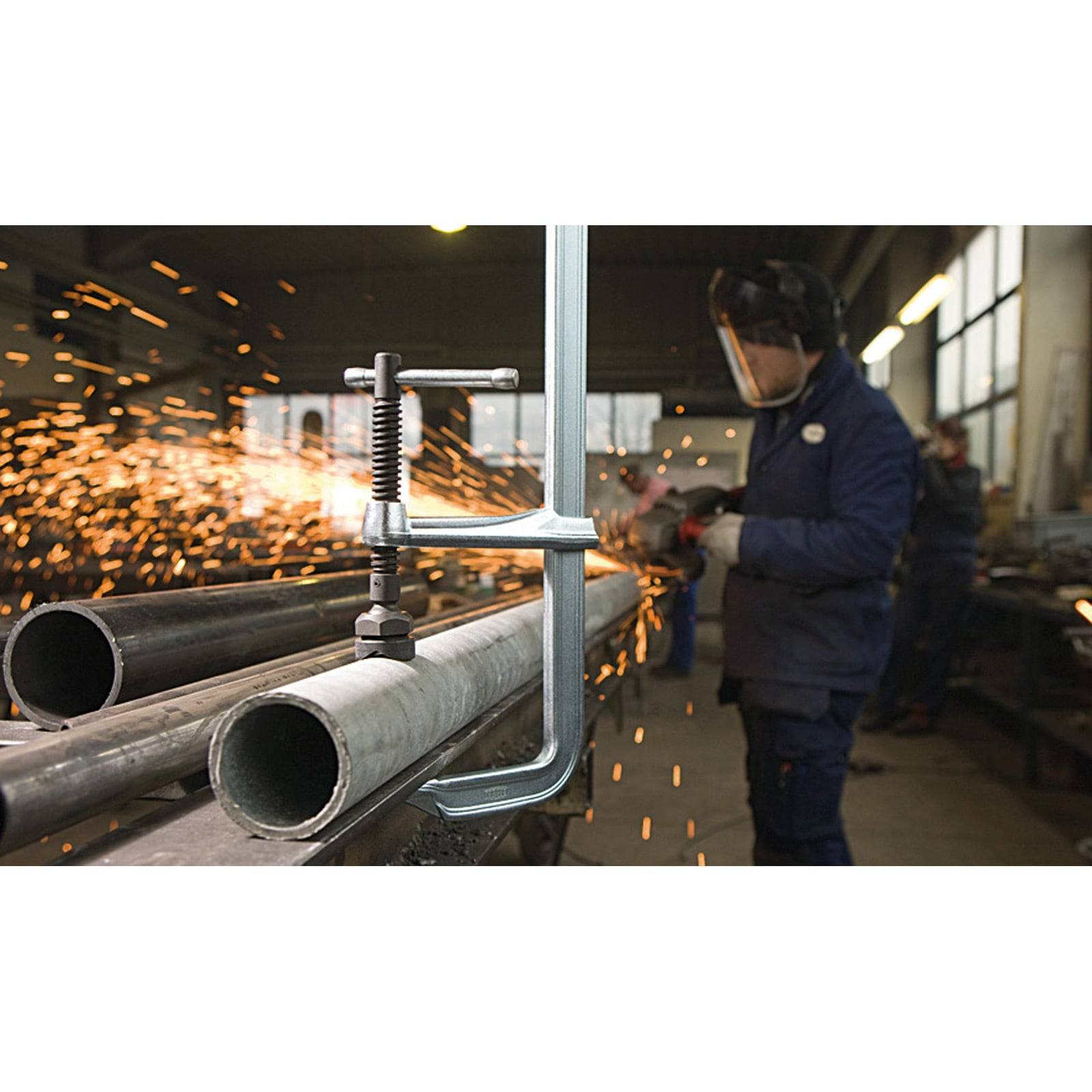 Ein Arbeiter in Schutzausrüstung schweißt Metallrohre in einer Werkstatt, umgeben von fliegenden Funken, was industrielle Handwerkskunst vermittelt.