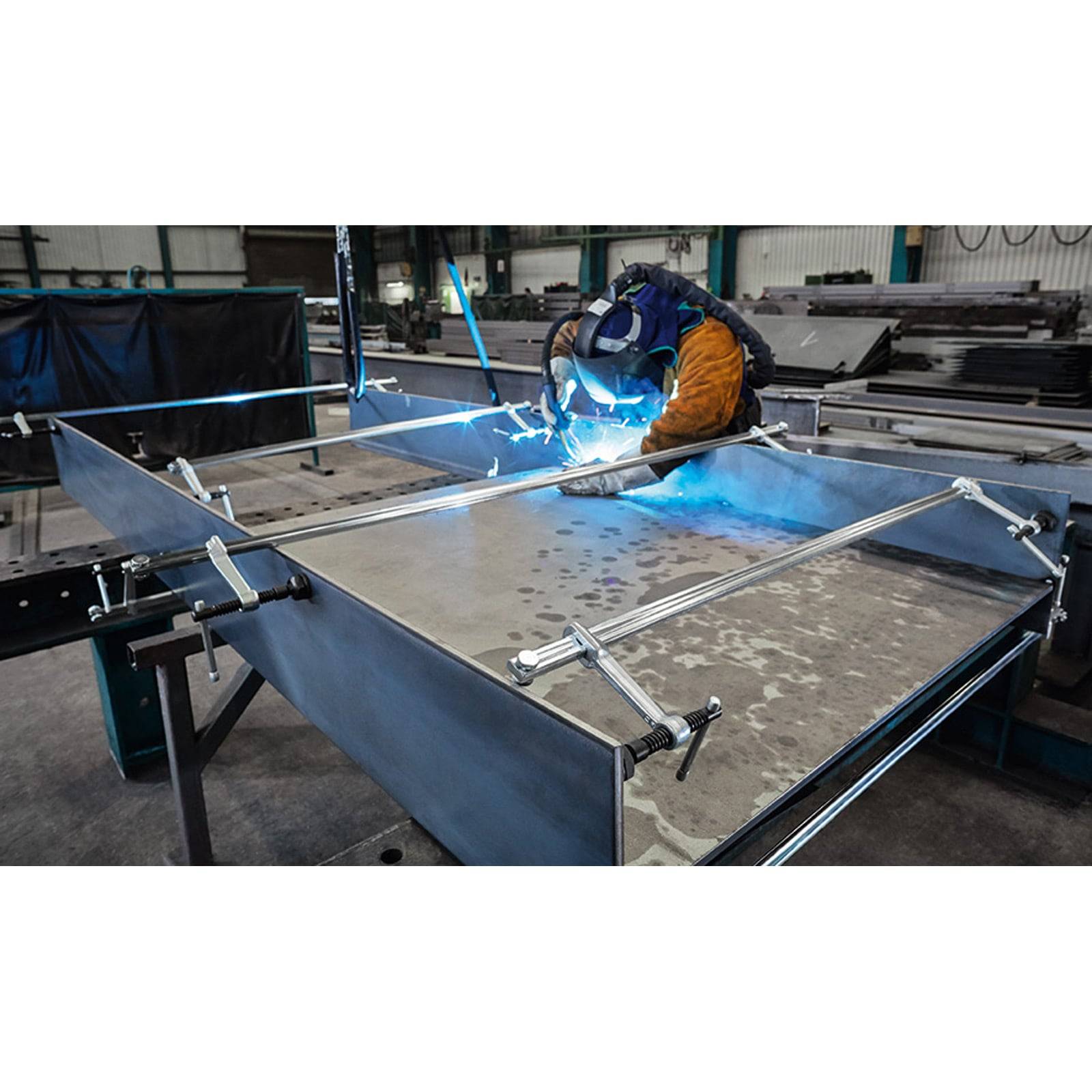 Ein Schweißer, der Schutzausrüstung trägt, schweißt ein großes Metallteil in einer Industrieumgebung. Blaue Funken und helles Licht sind sichtbar.