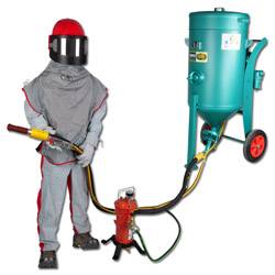 Eine Person in Schutzausrüstung bedient eine Sandstrahlmaschine, die über Schläuche mit einem unter Druck stehenden Behälter auf Rädern verbunden ist.