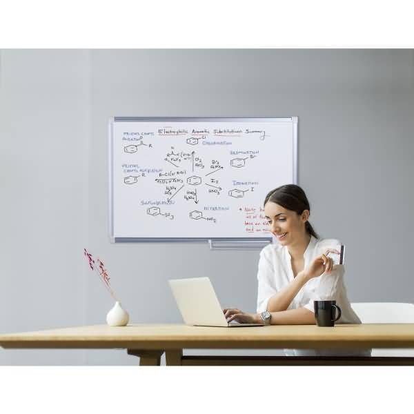 Eine Frau in einer weißen Bluse arbeitet an einem Laptop an einem Schreibtisch mit einer Tasse, vor einem Whiteboard, das mit handschriftlichen Chemie-Notizen und Diagrammen bedeckt ist.