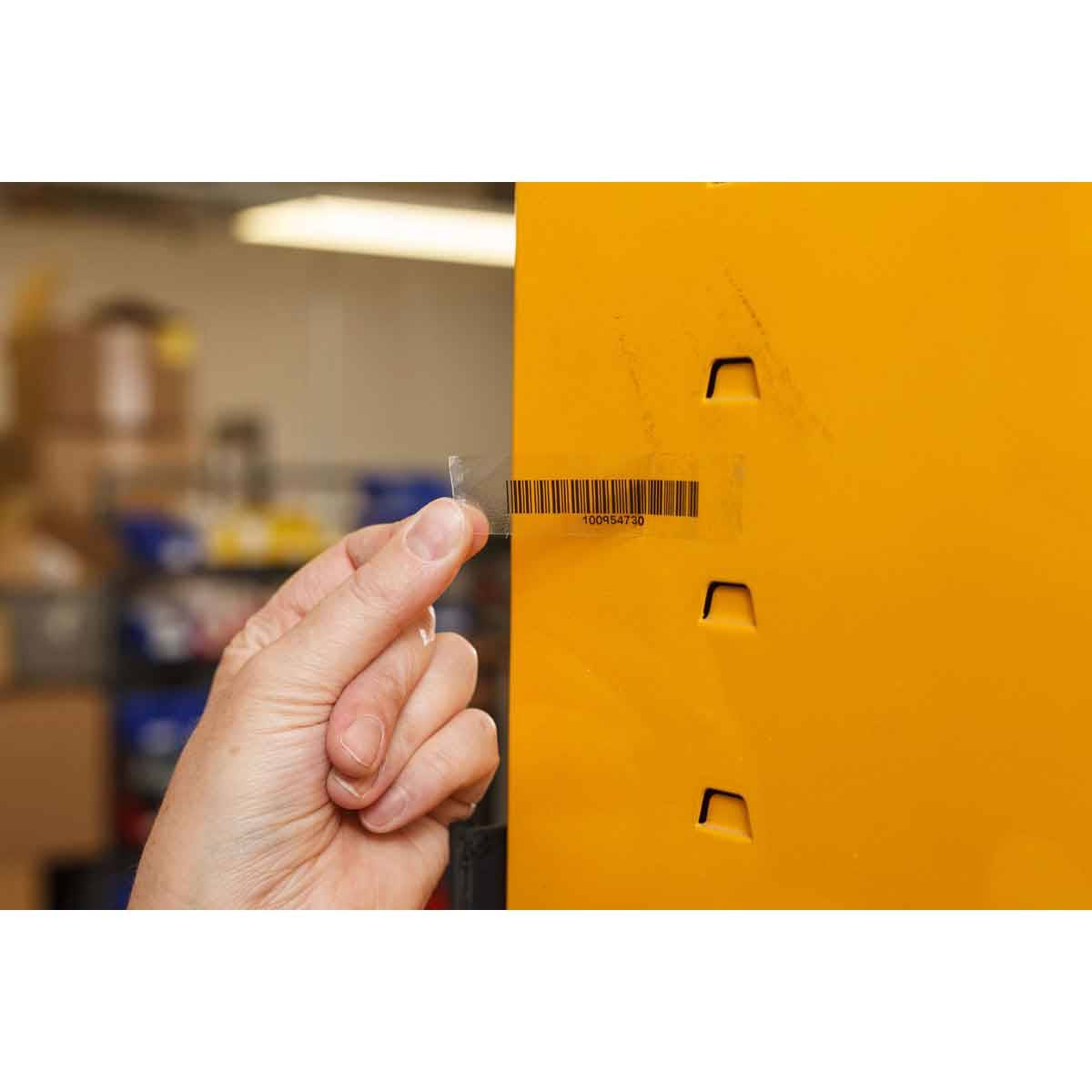 Das manuelle Abziehen eines durchsichtigen Sicherheitssiegels von einer gelben Maschine oder einem Schrank mit Barcode-Etikett; unscharfer Werkstatt- oder Lagerhallenhintergrund.