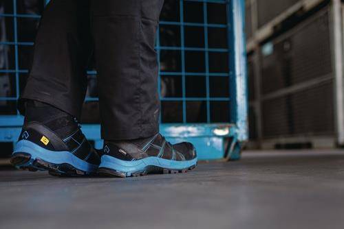 Person trägt schwarze Hose und blaue Wanderschuhe, steht drinnen in der Nähe eines Metallrostes, was auf eine industrielle oder Lagerhallen-Umgebung hindeutet.