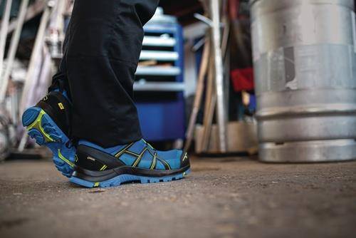 Eine Person trägt blaue und schwarze Wanderschuhe und steht in einer Werkstatt, mit verschwommenen Werkzeugen und Regalen im Hintergrund.