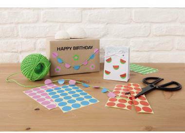 Geburtstagsgeschenkverpackungszubehör auf einem Tisch, einschließlich gemusterter Aufkleber, Schere, Schnur, ein grüner Wollknäuel und eine Geschenktüte mit der Aufschrift „ALLES GUTE ZUM GEBURTSTAG