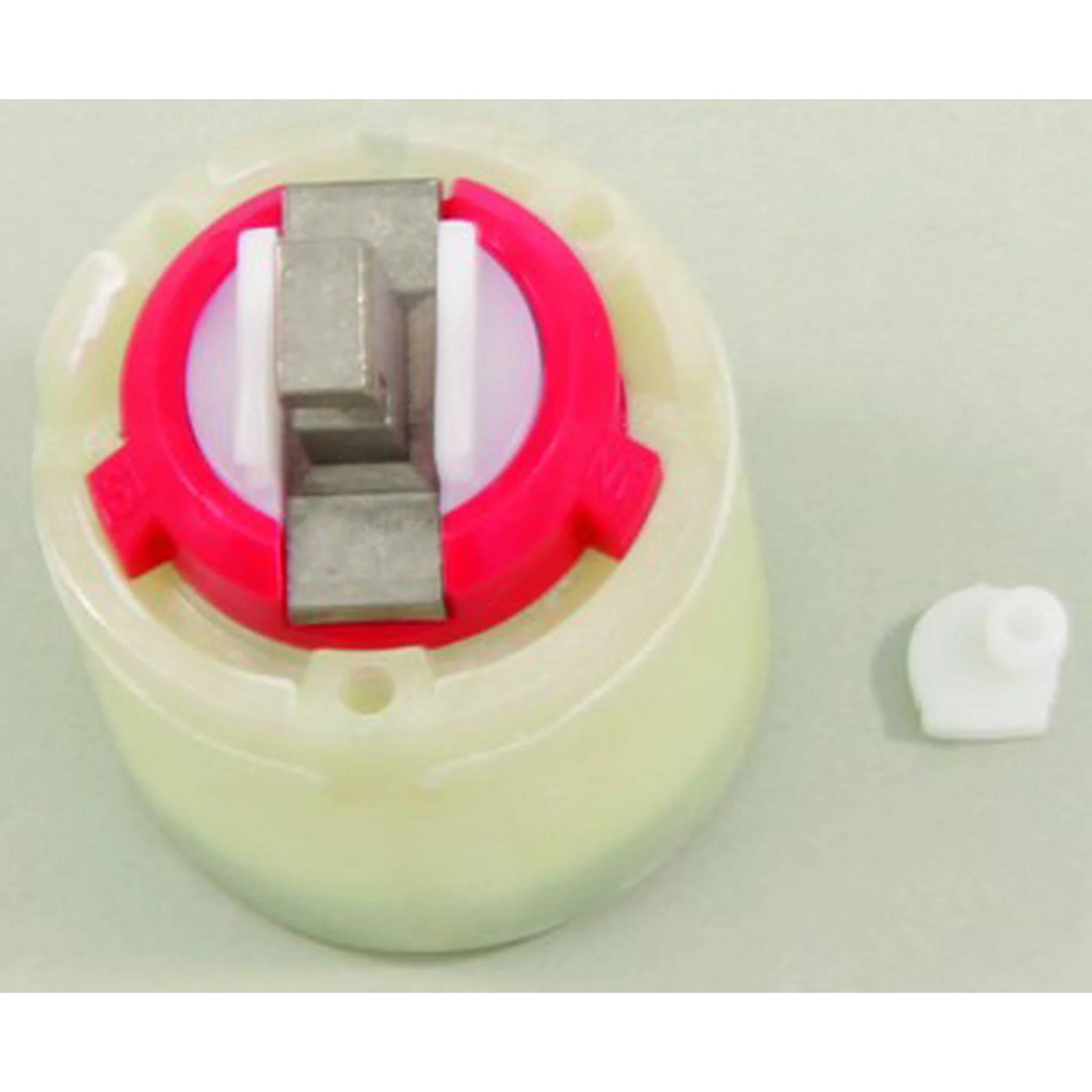 Kleine mechanische Baugruppe mit einer durchsichtigen zylindrischen Basis, rotem Ring und grauem Metallblock; ein separates weißes Kunststoffteil liegt rechts daneben.