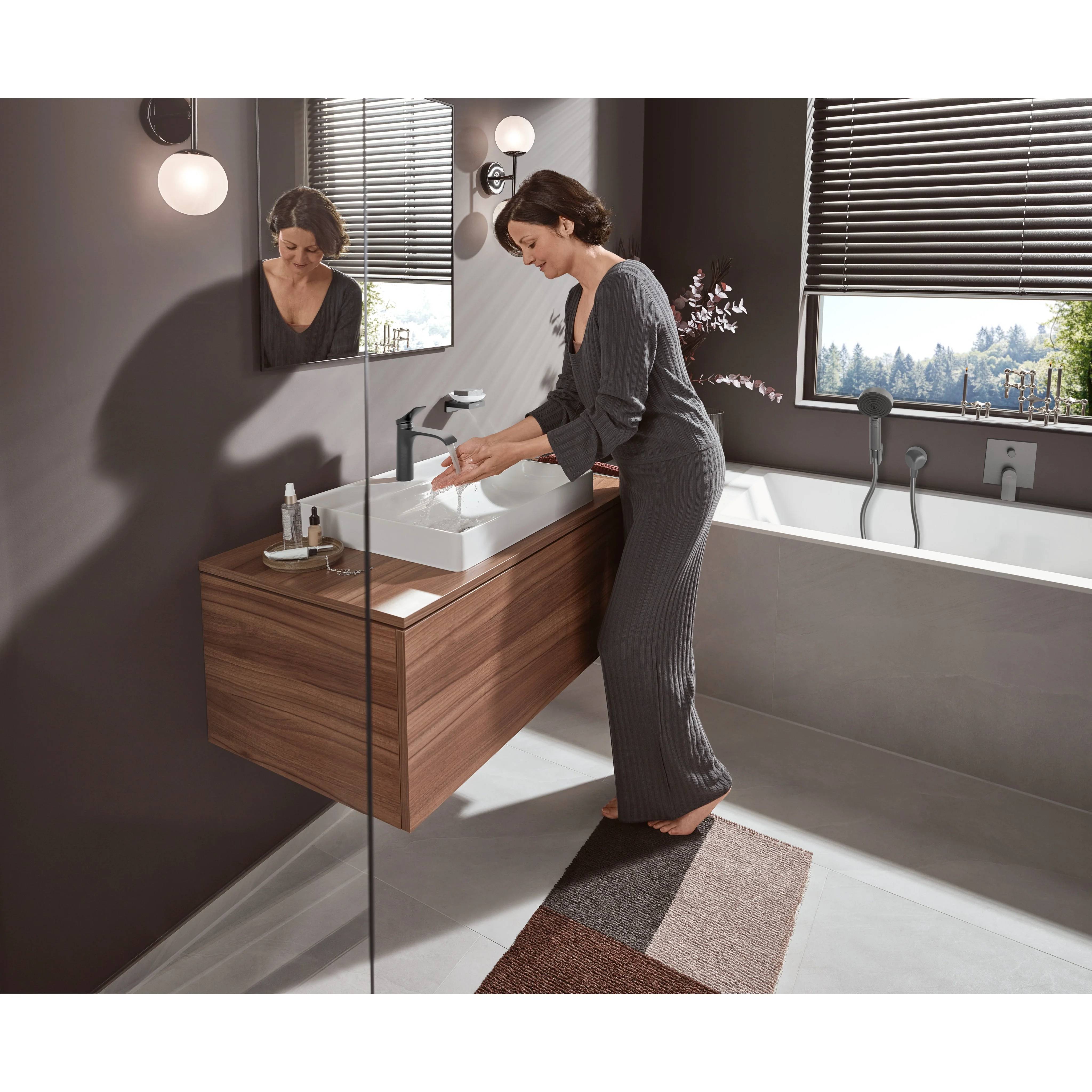 Frau wäscht sich die Hände an einem modernen wandmontierten Badezimmerbecken neben einer Badewanne und Fensterjalousien in einem zeitgenössischen Badezimmer.