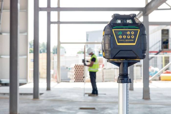 Eine Baustelle mit Fokus auf ein Lasernivelliergerät im Vordergrund; ein Arbeiter misst im unscharfen Hintergrund.