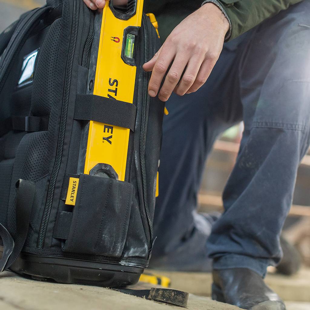 Eine Person, die eine gelbe Stanley-Wasserwaage in eine schwarze Transporttasche neben ihren Stiefeln auf einer Baustelle legt.