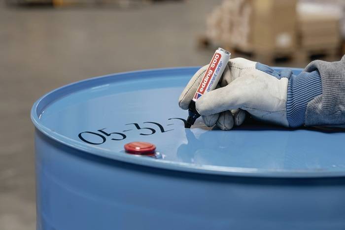 Eine behandschuhte Hand schreibt mit einem Marker '05-327' in Schwarz auf ein blaues Industriefass, was auf eine Kennzeichnung oder Organisation in einer Lagerhalle hinweist.