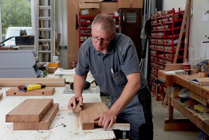 Ein Mann in einer Werkstatt schleift einen Holzblock auf einem Tisch, umgeben von Holzstücken und Werkzeugen, und konzentriert sich dabei sehr auf seine Aufgabe.
