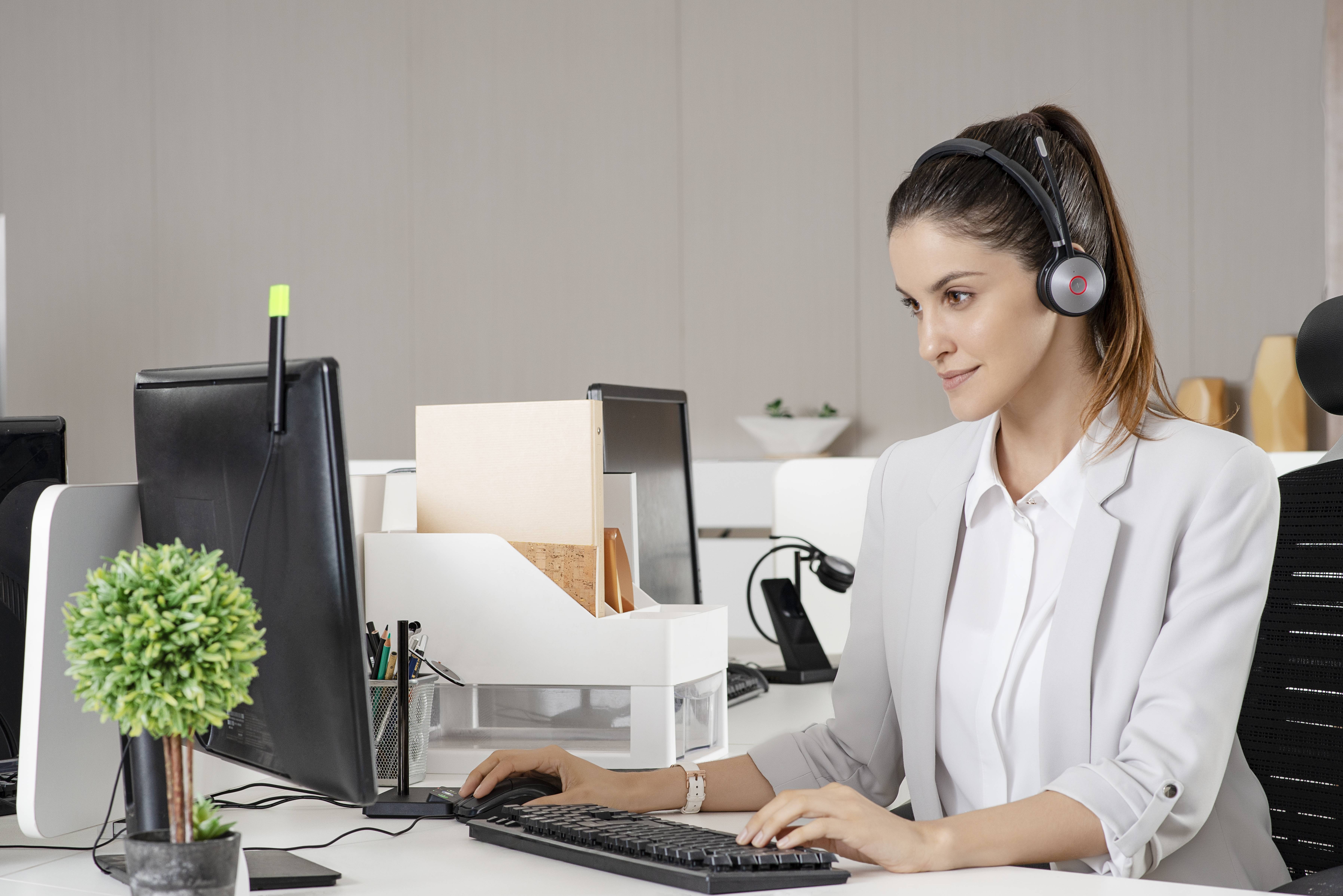 Eine Frau mit einem Headset arbeitet an einem Desktop-Computer in einem Büro, was auf Kundenbetreuung oder Call-Center-Arbeit hindeutet.