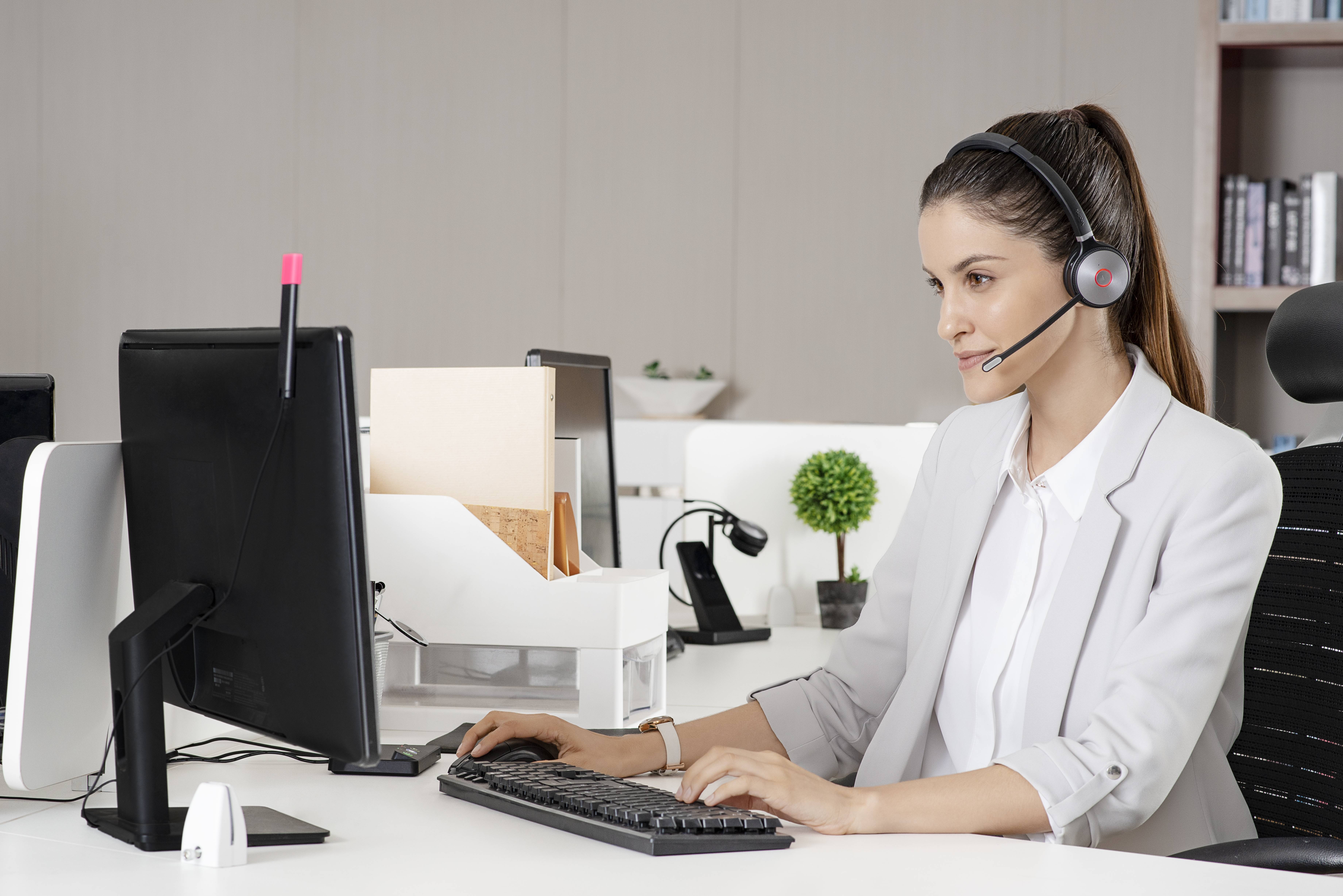 Frau mit Kopfhörer arbeitet an einem Desktop-Computer in einem modernen Büro, tippt und spricht als Kundendienstmitarbeiterin.