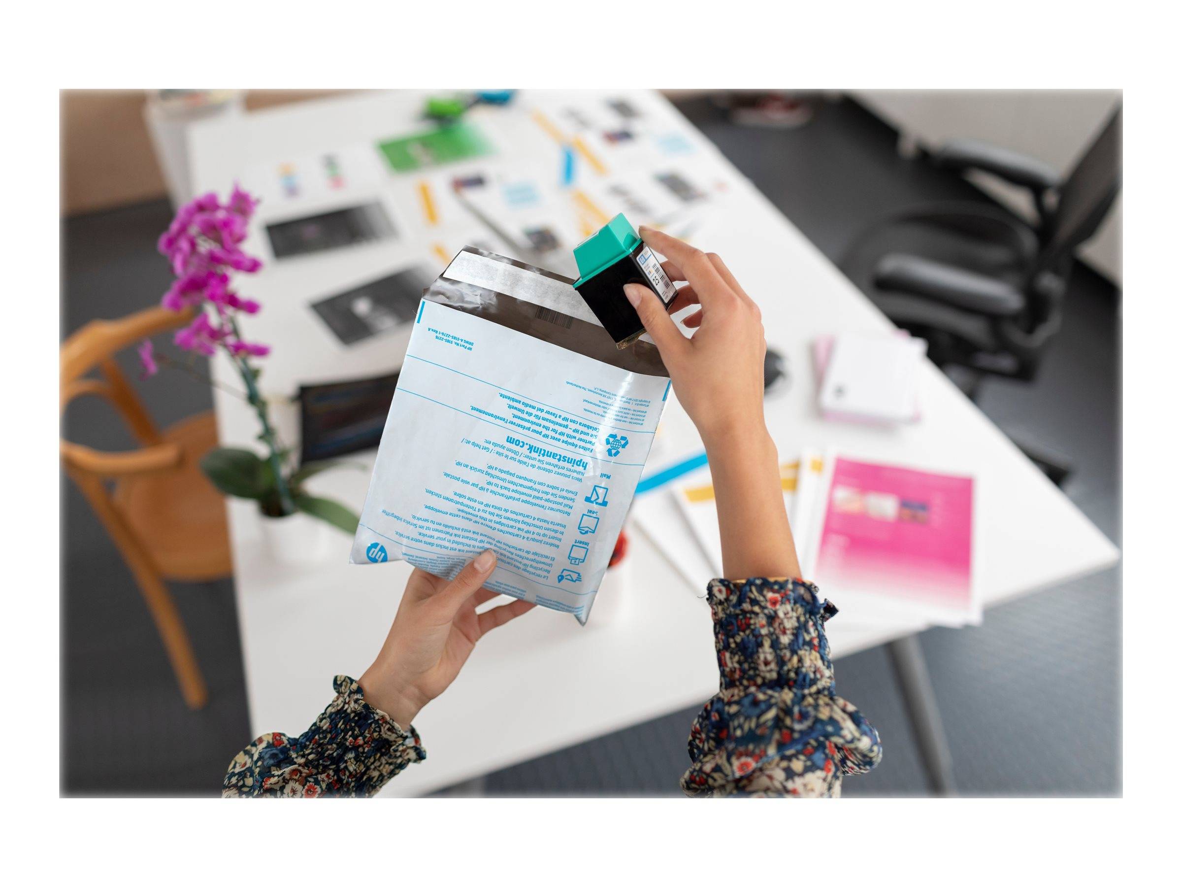 Hände packen ein kleines Paket oder Päckchen über einem Bürotisch aus, mit einer rosa Orchidee und unscharfen Arbeitsmaterialien im Hintergrund.