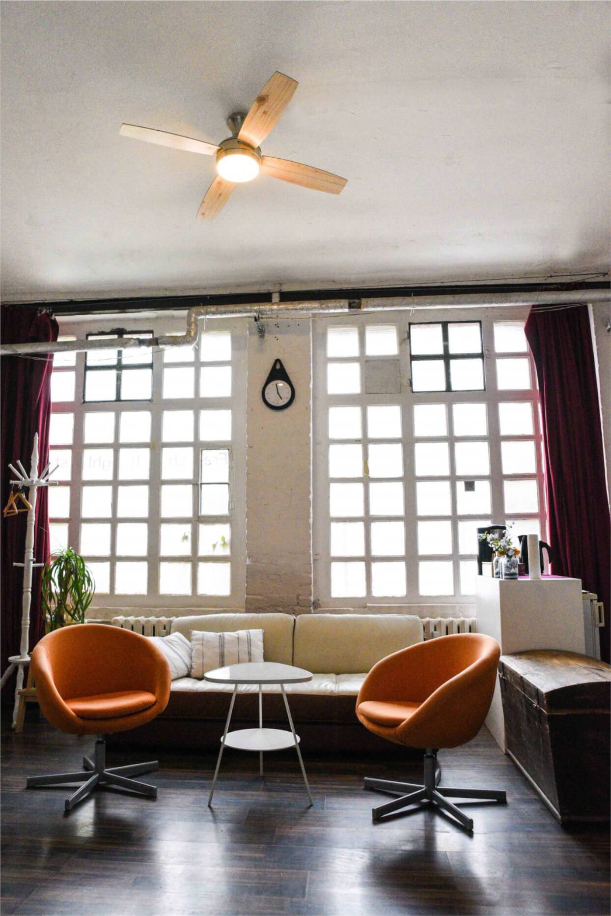 Ein gemütliches Zimmer mit großen Sprossenfenstern, einer Wanduhr und drei modernen Stühlen um einen kleinen Tisch. Rote Vorhänge und eine Topfpflanze schaffen Wärme.