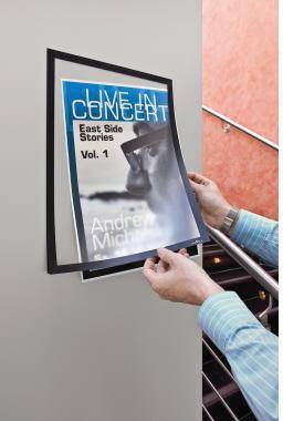 Hände platzieren ein gerahmtes Poster an einer Wand. Das Poster zeigt die Aufschriften „LIVE IN CONCERT