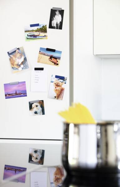 Fotoabdrucke und Schnappschüsse mit Klebeband auf einem weißen Kühlschrank befestigt, mit einem unscharfen Metalltopf im Vordergrund.