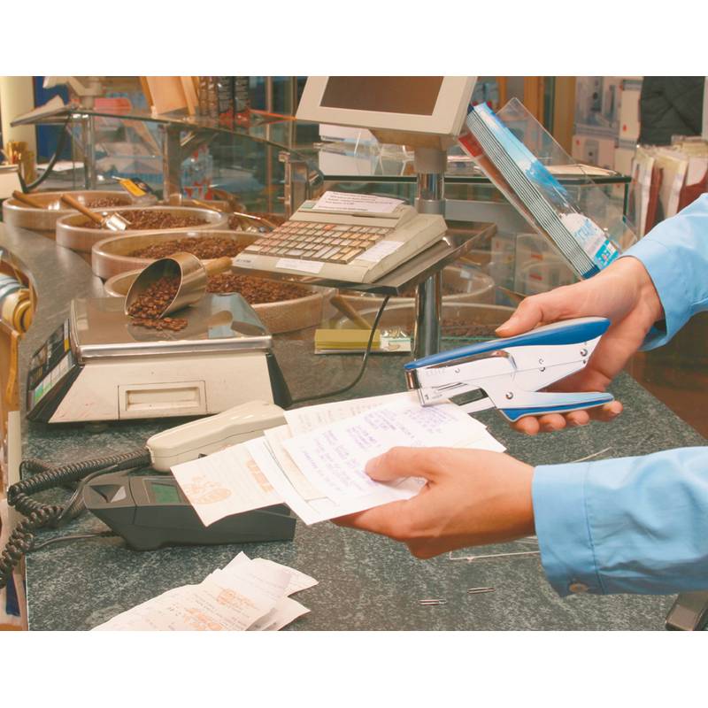 Eine Person verwendet einen blauen Hefter, um Quittungen an einem Verkaufstresen mit einer Registrierkasse und Kaffeebohnen in Tabletts in der Nähe zusammenzuheften.