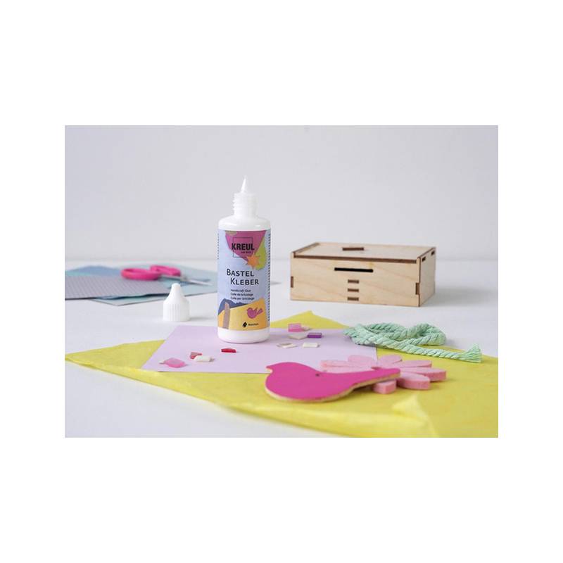 Bastelstation mit einer Klebeflasche, einer Holzkiste, rosa und gelben Papierformen, einer Schere und einem Band auf einem Tisch.